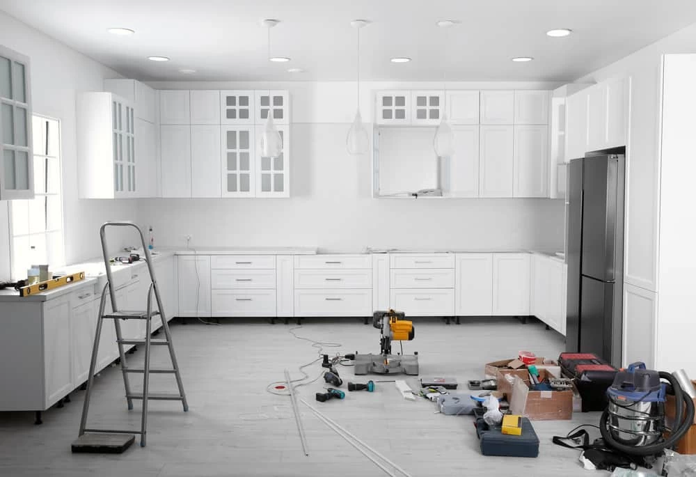 A modern kitchen under renovation, featuring white cabinets, tools scattered on the floor, a ladder, and a countertop saw in the center. This bustling scene embodies the essence of a dedicated home remodeler at work.