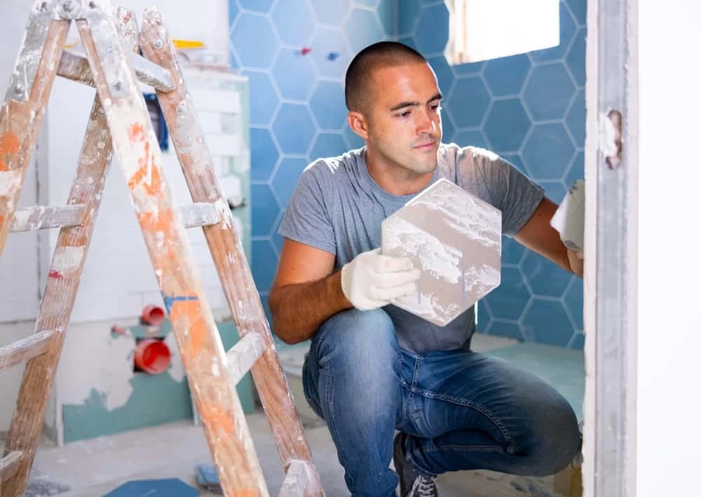 A man in a gray shirt and jeans is meticulously installing hexagonal tiles on a bathroom wall, emblematic of a detailed home remodeling project. Nearby, a ladder with paint splatters hints at ongoing work across the space.