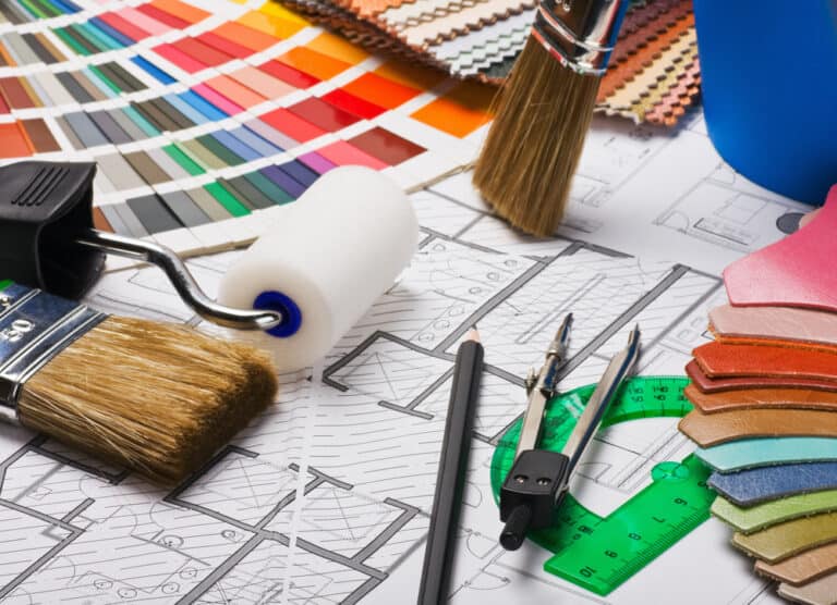 A variety of painting tools, color swatches, brushes, a roller, pencils, and a protractor are placed on top of architectural blueprints for a kitchen renovation.