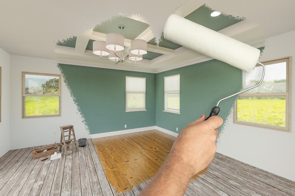 A home remodeler’s hand holds a paint roller, transforming a beige room to vivid green. The space features wooden floors, a modern chandelier, and two large windows showcasing lush greenery.