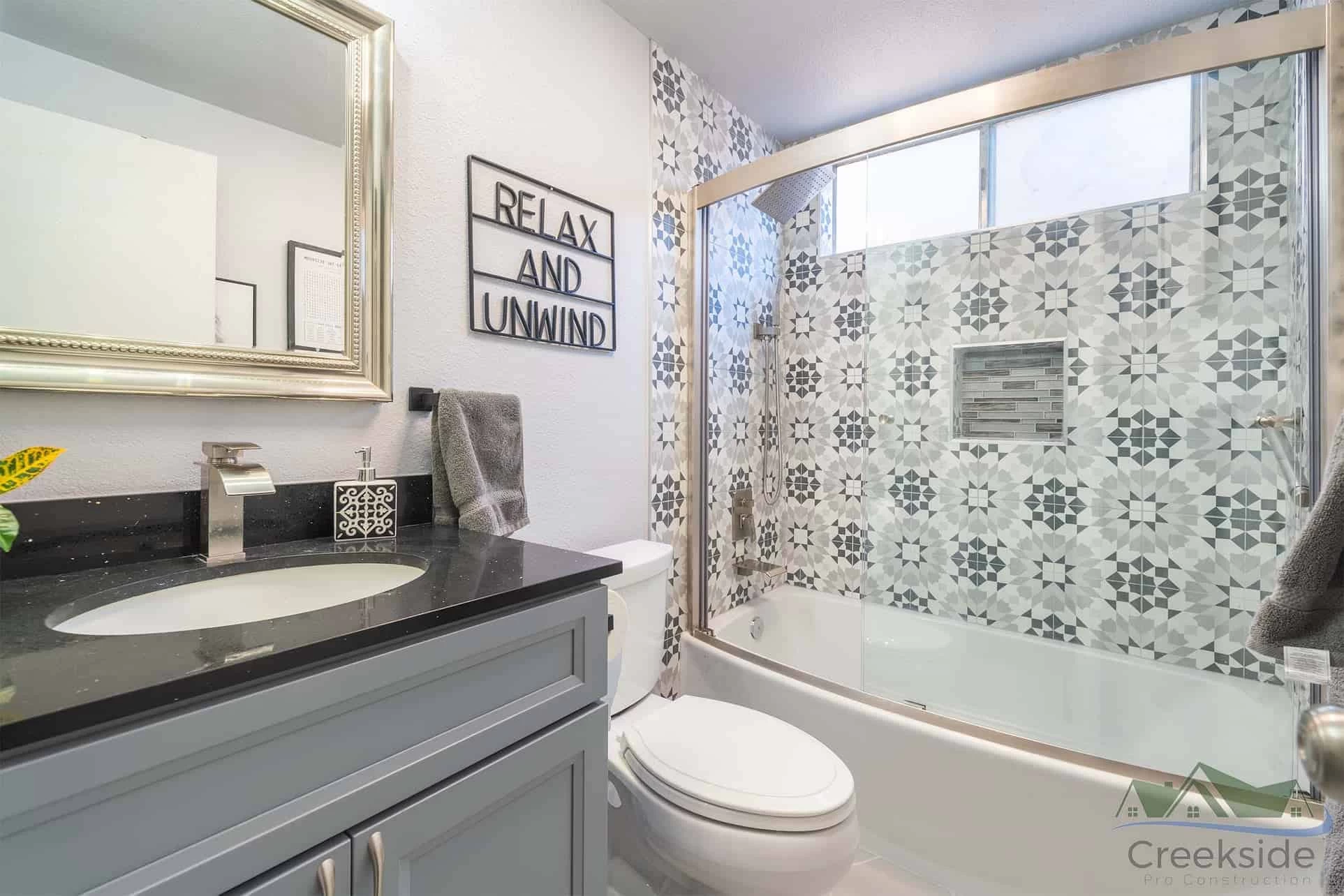 A modern bathroom with a white sink, black countertop, and floral-patterned tiles on the wall above a bathtub with a glass screen. Sign reads "RELAX AND UNWIND." Perfect inspiration for anyone considering home remodeling or seeking ideas from their trusted kitchen remodeler.