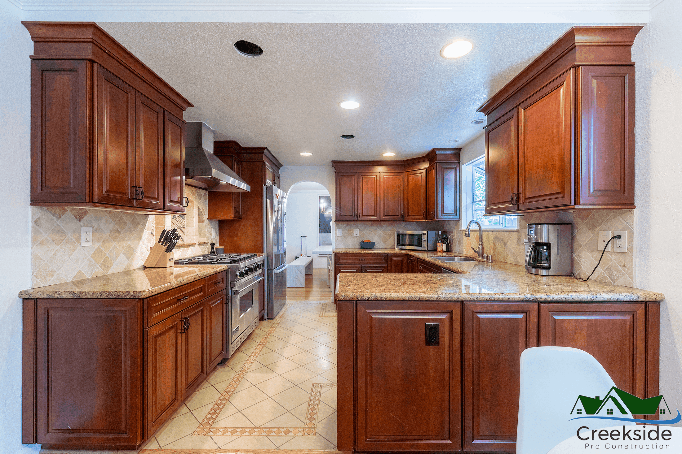 A spacious kitchen with dark wood cabinets, granite countertops, stainless steel appliances, and beige tile flooring. Perfect for those considering a kitchen remodeling project. Creekside Pro Construction logo. A spacious kitchen with dark wood cabinets, granite countertops, stainless steel appliances, and beige tile flooring.