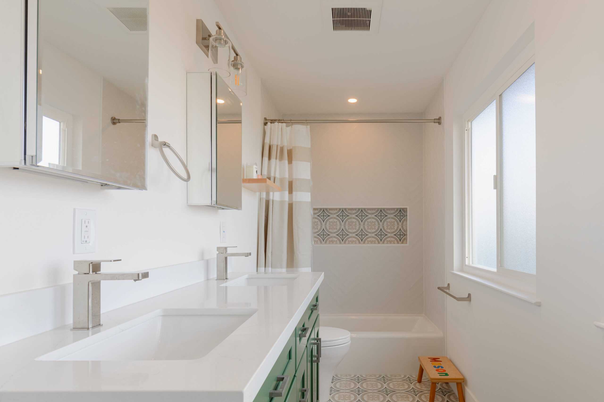 A modern bathroom with dual sinks, green cabinets, a bathtub with a patterned tile accent, a striped shower curtain, and a window showcases the craftsmanship of our dedicated home remodeler.