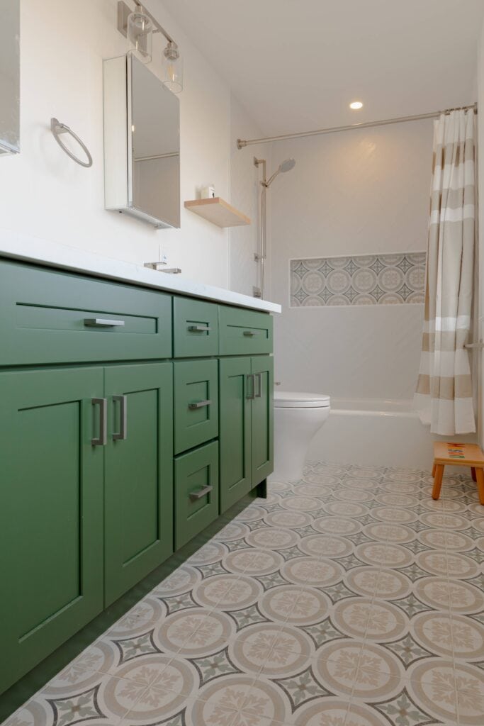 Modern bathroom with green vanity and patterned floor tiles, part of a recent home renovation. White walls, mirrored cabinet, and shower with a beige and white striped curtain complete the stylish look.