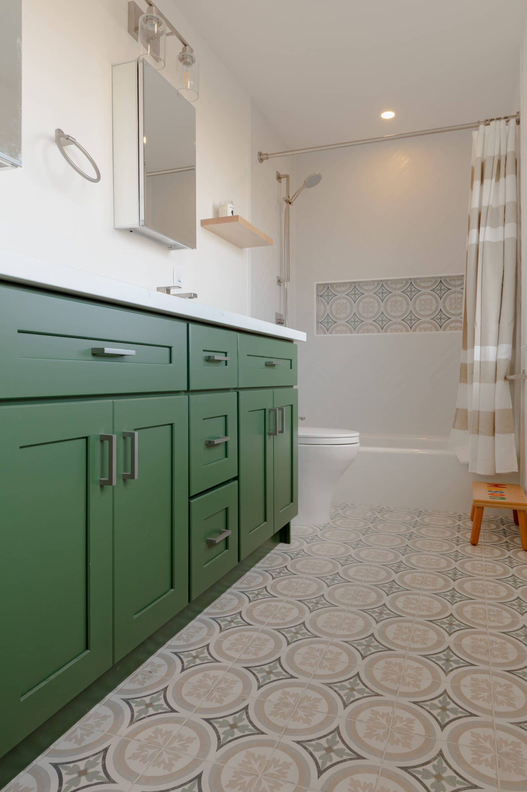 Modern bathroom with green vanity and patterned floor tiles, part of a recent home renovation. White walls, mirrored cabinet, and shower with a beige and white striped curtain complete the stylish look.