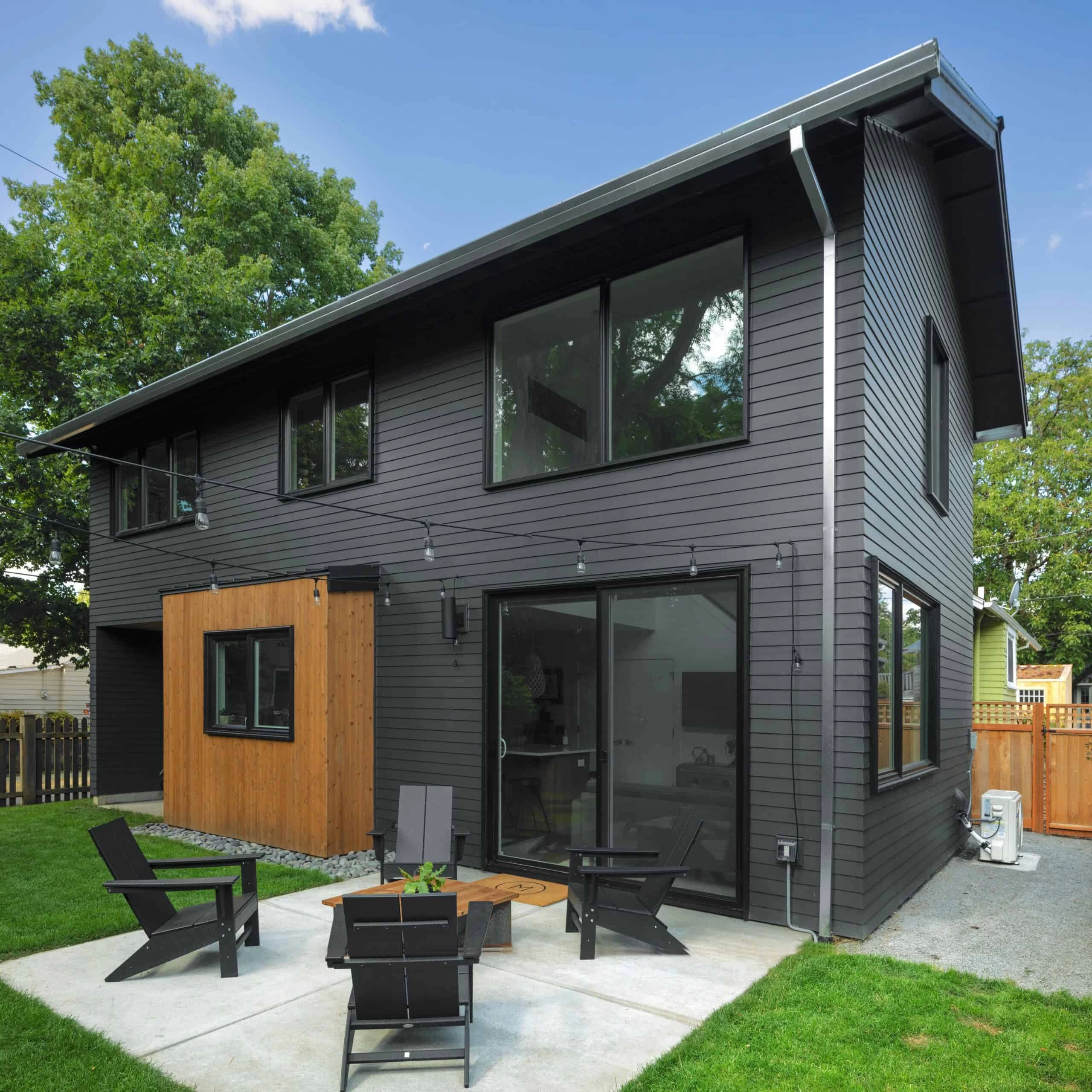 A modern two-story house with dark grey siding, large windows, a wooden accent wall, and a small patio with black chairs and a fire pit.