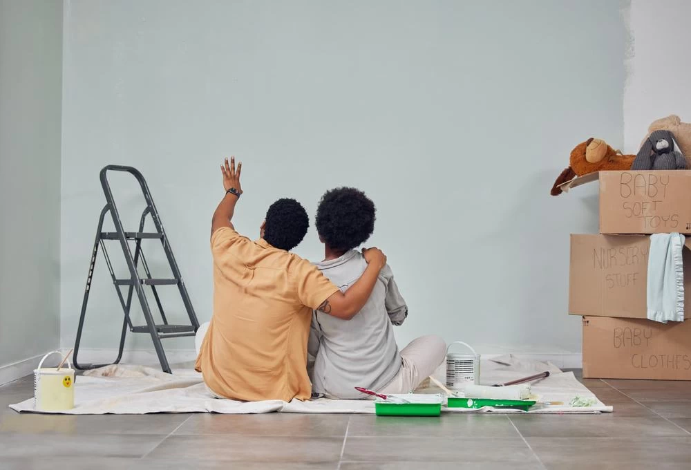 Two people sit on the floor, hugging and looking at a freshly painted wall, surrounded by painting supplies and moving boxes—a true scene of home remodeling success.