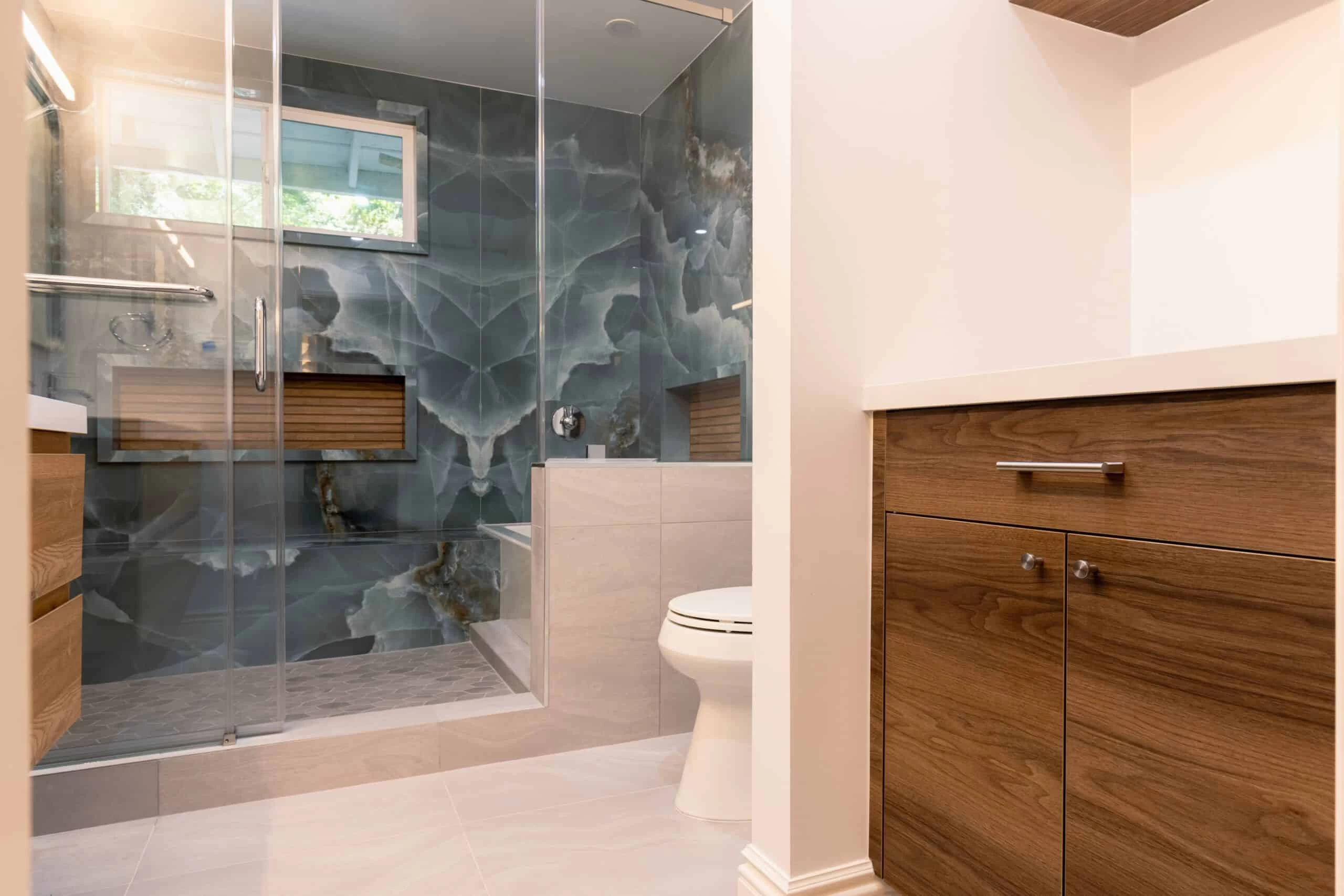 Modern Bath Room Design – Contra Costa County, CA Modern bathroom with a glass-enclosed shower, blue marble accent wall, wooden cabinets, and a white toilet on light-colored tiles.