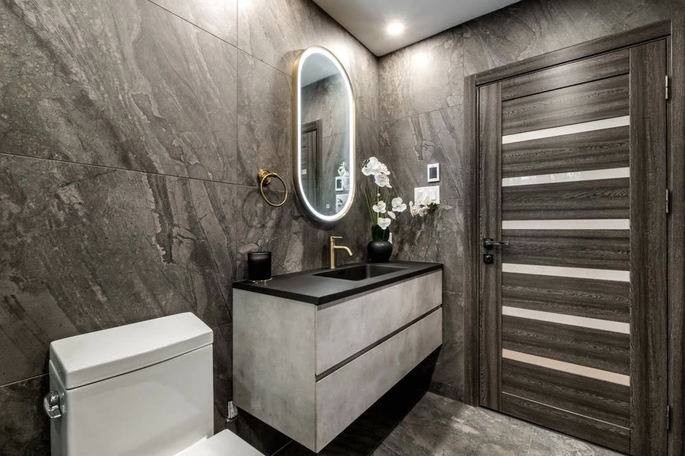 Modern bathroom with dark tile walls, oval mirror with backlighting, floating vanity with black sink, toilet, and a wooden door with horizontal accents. Perfect for any home remodeler seeking a sleek design in Contra Costa County, CA.