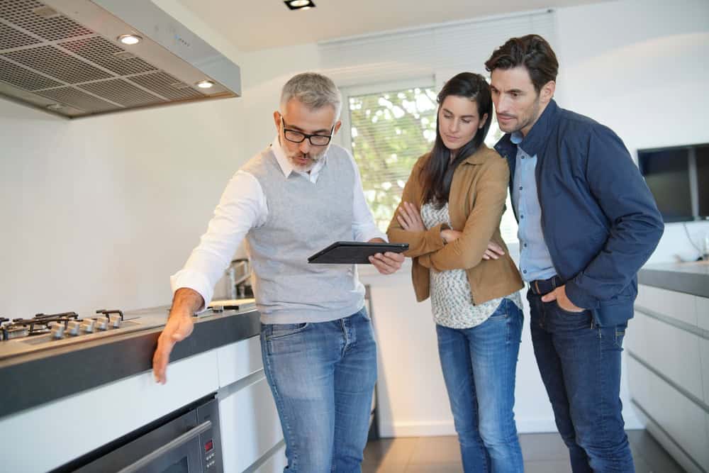 A salesperson holding a tablet explains the features of a kitchen appliance to a couple planning a kitchen remodeling in their modern kitchen.