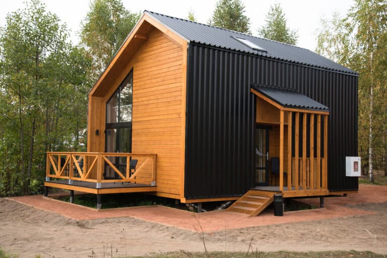 Modern tiny house with a wooden exterior and dark metal siding, featuring a small porch with railing, set amidst a wooded area. Perfect for those considering home remodeling or the expertise of a skilled home remodeler to transform your space into a cozy woodland retreat.