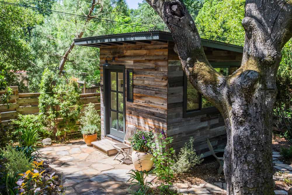 A small, rustic wooden cabin with large windows sits in a lush garden surrounded by trees, plants, and a stone pathway, perfect for those dreaming of home remodeling projects or envisioning a charming kitchen renovation.