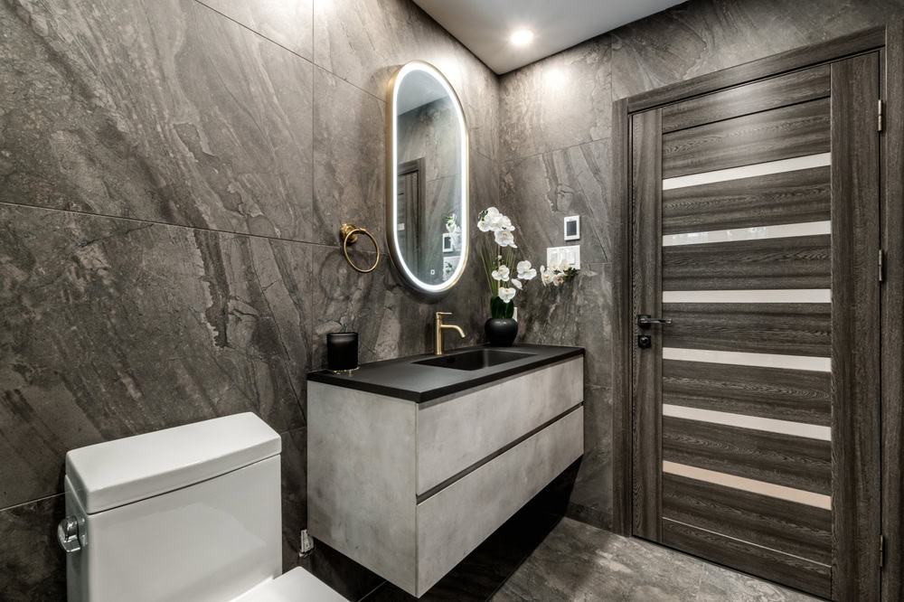 Modern bathroom with dark tile walls, oval mirror with backlighting, floating vanity with black sink, toilet, and a wooden door with horizontal accents. Perfect for any home remodeler seeking a sleek design.