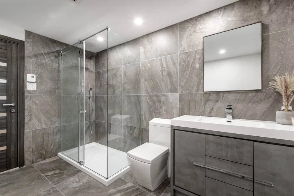 Modern bathroom with gray tile walls and floor, walk-in glass shower, white toilet, vanity with rectangular mirror, and a small potted plant in Contra Costa County, CA.