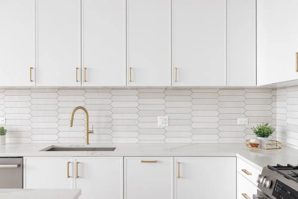 Modern kitchen with white cabinets, brass hardware, white geometric tile backsplash, stainless steel sink with brass faucet, and small plants. Ideal inspiration for anyone planning a home renovation or considering a professional kitchen remodeler.