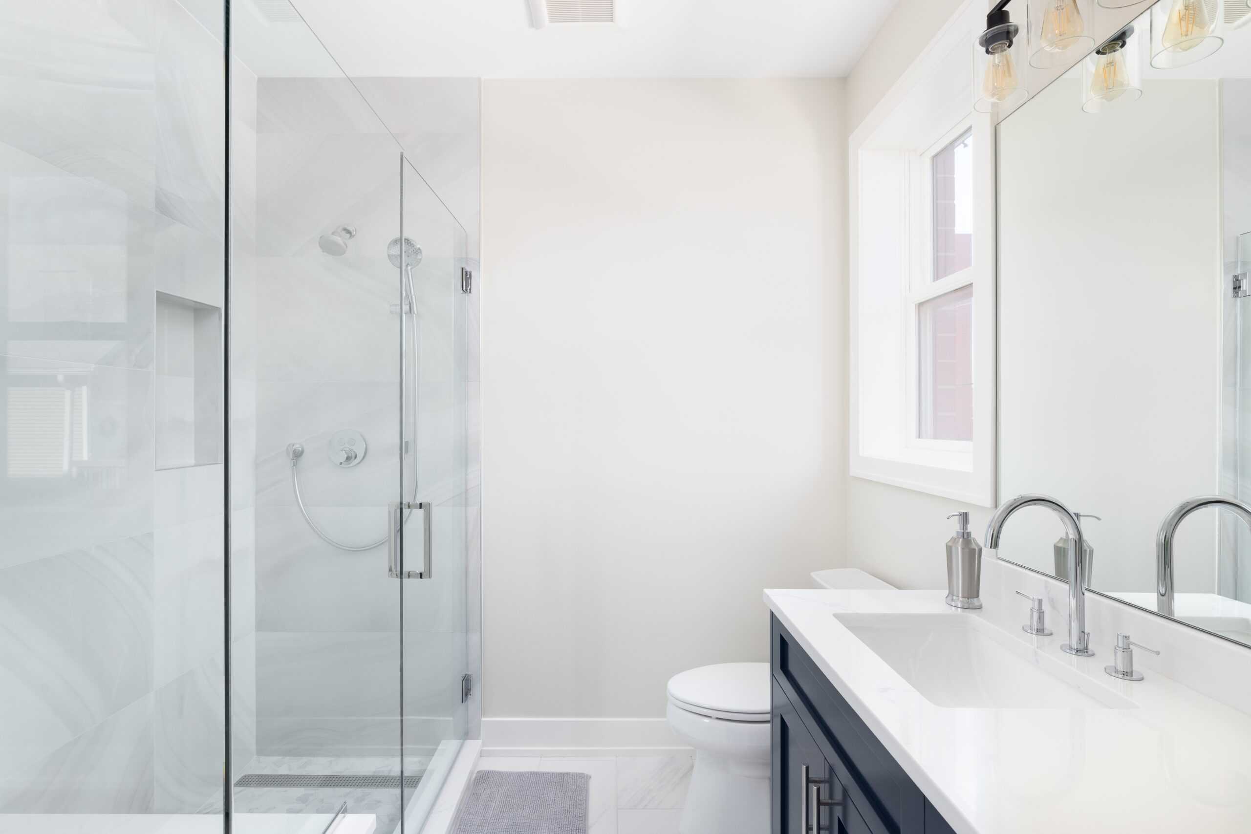 Modern bathroom with glass-enclosed shower, white walls, and vanity with dual sinks and mirrors. Bright with natural light from the window, it's a seamless blend of elegance and functionality, just like a sophisticated kitchen renovation.