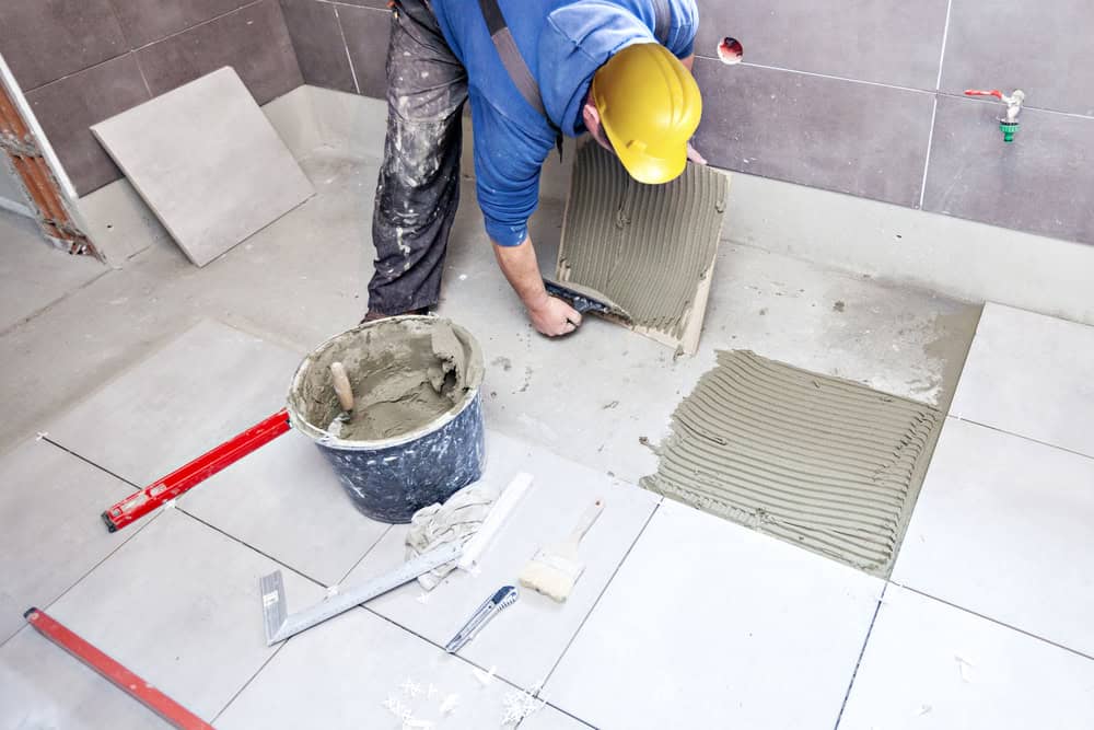 A construction worker in a yellow hard hat applies adhesive to the floor while installing large, gray tiles during a home remodeling project.