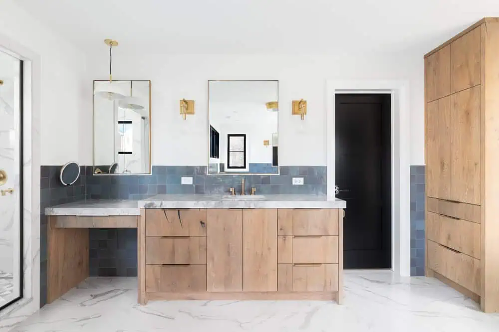 Modern bathroom with wooden cabinets, dual mirrors, two sinks, marble countertops, blue-tiled backsplash, gold fixtures, and white marbled flooring—perfect for anyone considering a home renovation project.